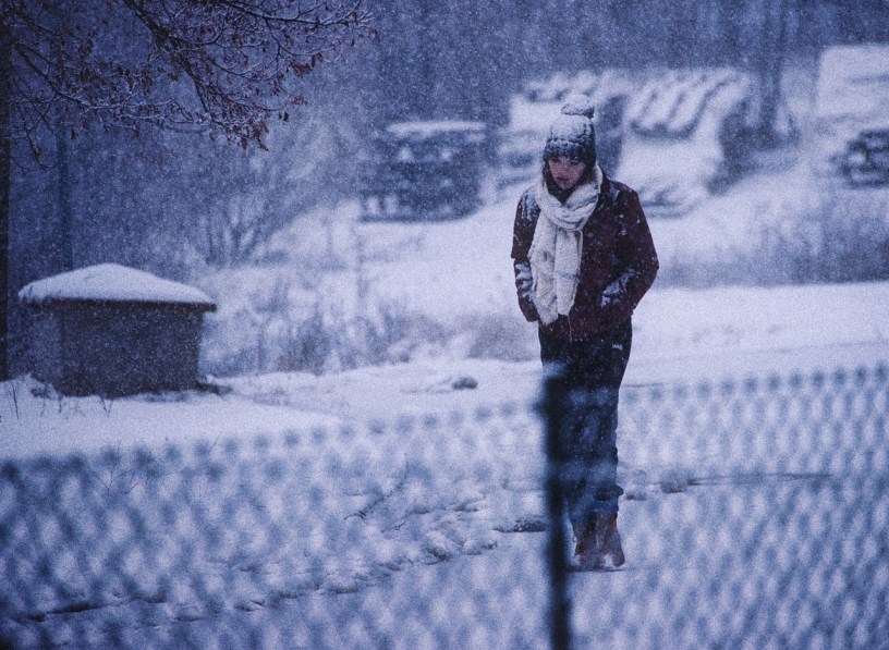 a girl walks along a chain-link fence during a winter blizzard