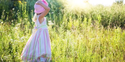 A little girl in a pastel striped dress and pink hat stands in a field looking toward the low-lying sun