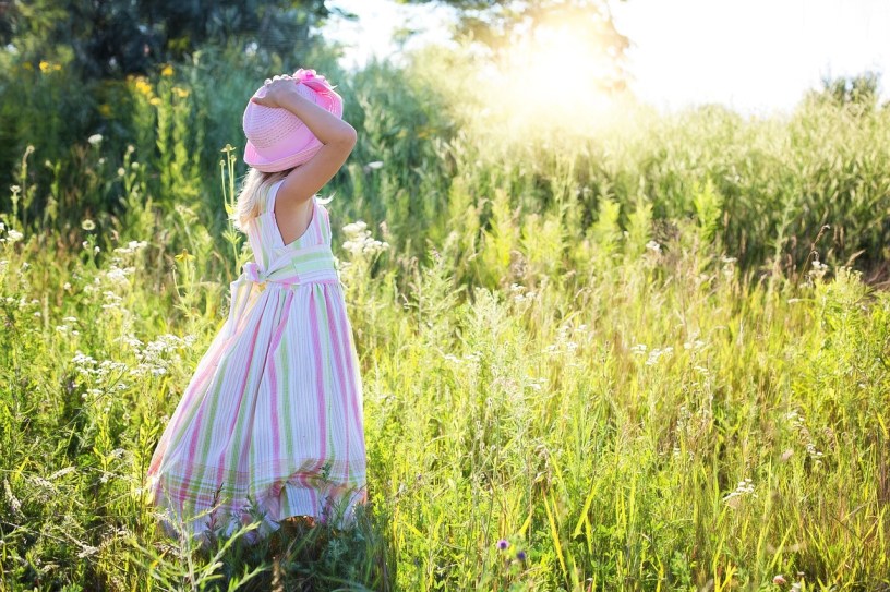 A little girl in a pastel striped dress and pink hat stands in a field looking toward the low-lying sun