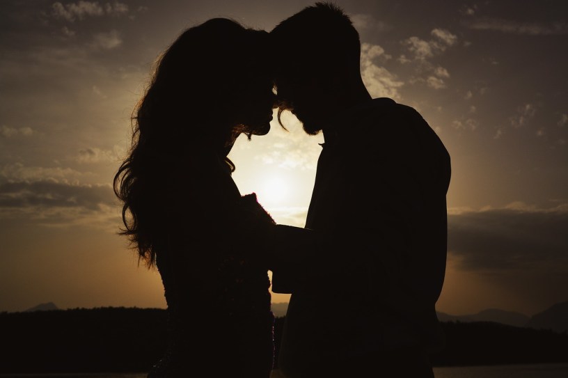 silhouette of a couple embracing, about to kiss, backed by the sun low in a partly cloudy sky