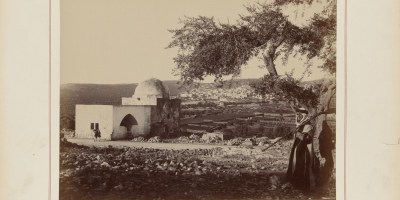 white exterior of Rachel’s Tomb, with its recognizable dome, is seen on the left, while the edifices of the city of Bethlehem grace hills in the background. The figures of traditionally dressed local inhabitants stand by the large olive tree