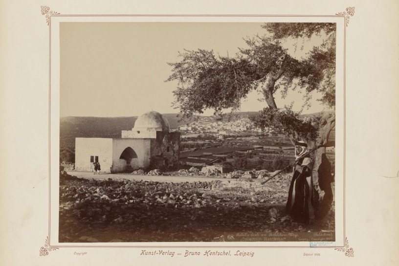 white exterior of Rachel’s Tomb, with its recognizable dome, is seen on the left, while the edifices of the city of Bethlehem grace hills in the background. The figures of traditionally dressed local inhabitants stand by the large olive tree