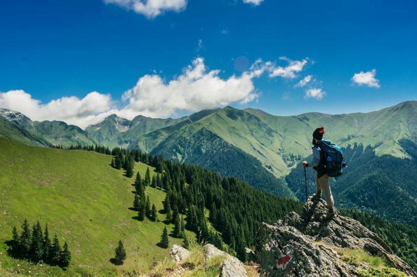 hiker on a rock outcropping overlooking a grassy slope ascending to a green mountain range