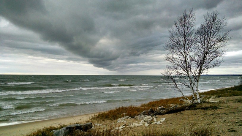 A low, dark gray cloud bank rolls over the north coast of Lake Michigan, as seen from the shore of the Upper Peninsula