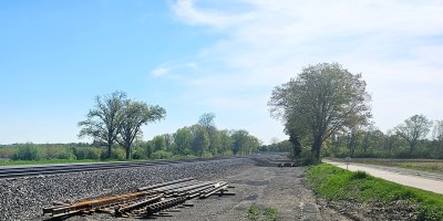 train tracks, with discarded pieces lying on the side, run off toward a partly cloudy horizon in a rural setting near a dirt road and trees