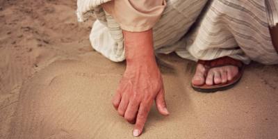 A hand writing in the sand, belonging to a person in a robe and sandals like Jesus in the Gospel of John