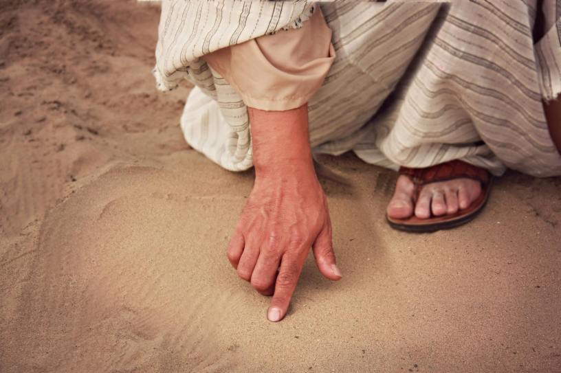 A hand writing in the sand, belonging to a person in a robe and sandals like Jesus in the Gospel of John