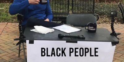 This is a manipulated photo of a classic Steven Crowder meme. Steven Crowder's face has been replaced with the wild, slightly confused expression of "Grandpa Joe". Grandpa Joe sits at a table in a public space with a sign that says, "Black people are cursed - Change my mind"